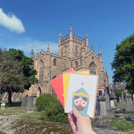 Set of 3 brightly coloured Robert the Bruce greeting cards are held up  in front of Dunfermline Abbey.
