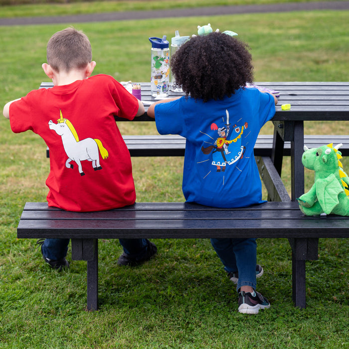 Children sit at a bench drawing. One wears a red tee featuring a unicorn and the other wears a blue tee with a knight.