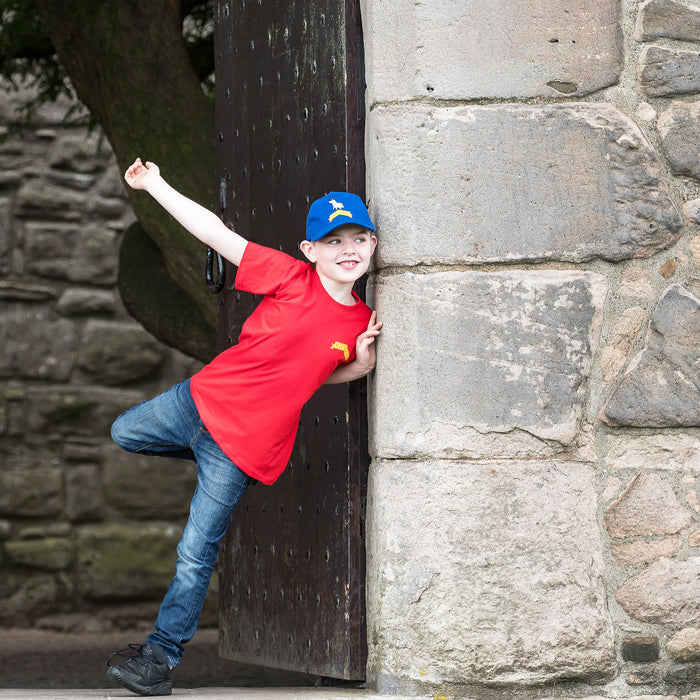 Child standing in a castle doorway is wearing the blue Unicorn cap and the Red Unicorn Tee.