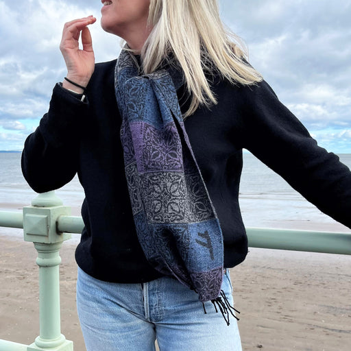 Person with blonde hair wears the purple and blue Runes scarf against a sea and beach background