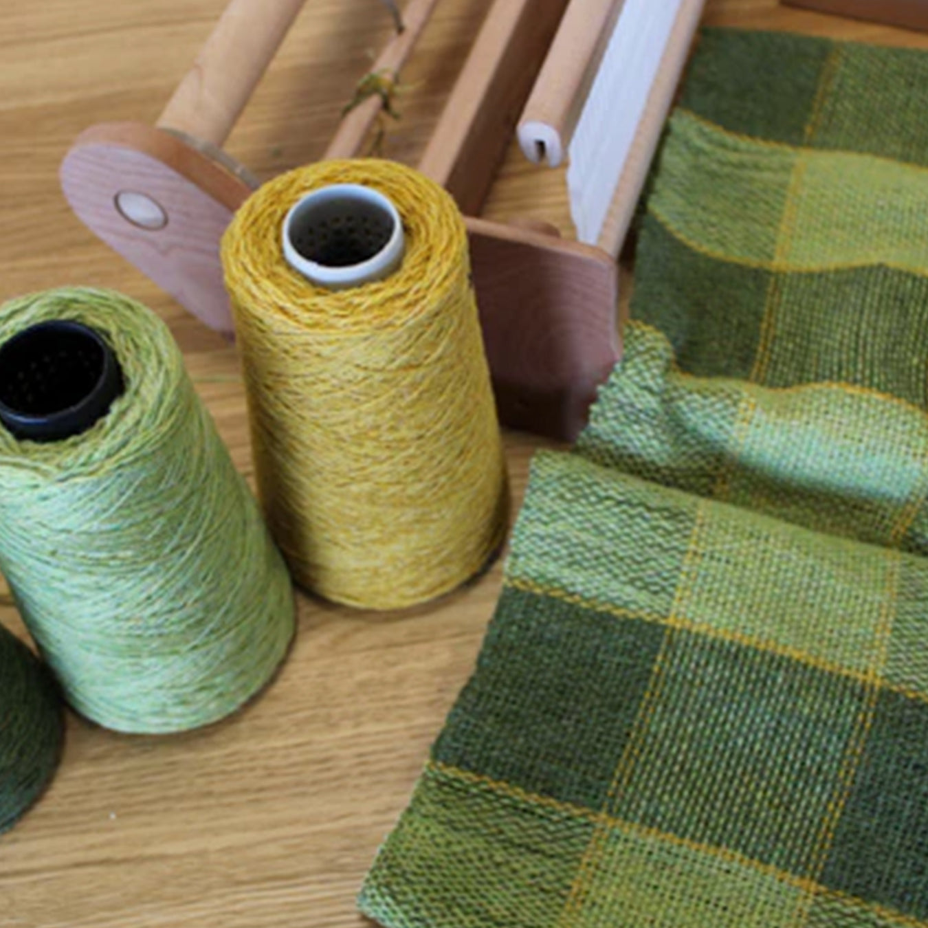 Yarns of yellow and green wool are placed on a wooden table next to some tartan weaved of the same yarns.