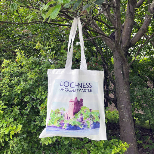 A white tote bag hangs from a tree branch. The tote features a colourful print of Urquhart Castle on the banks of Loch Ness. 