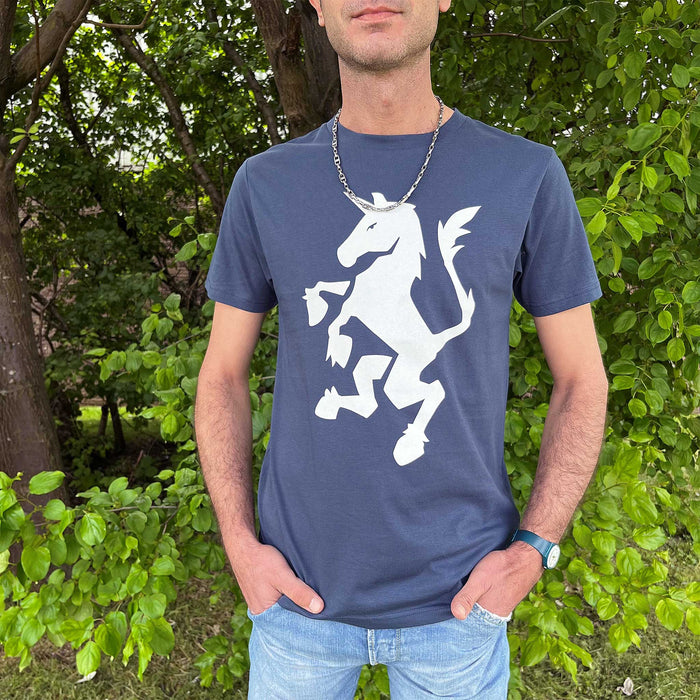 Person standing in front of some trees and greenery wears light blue jeans with a navy t-shirt which features a white standing unicorn print. 