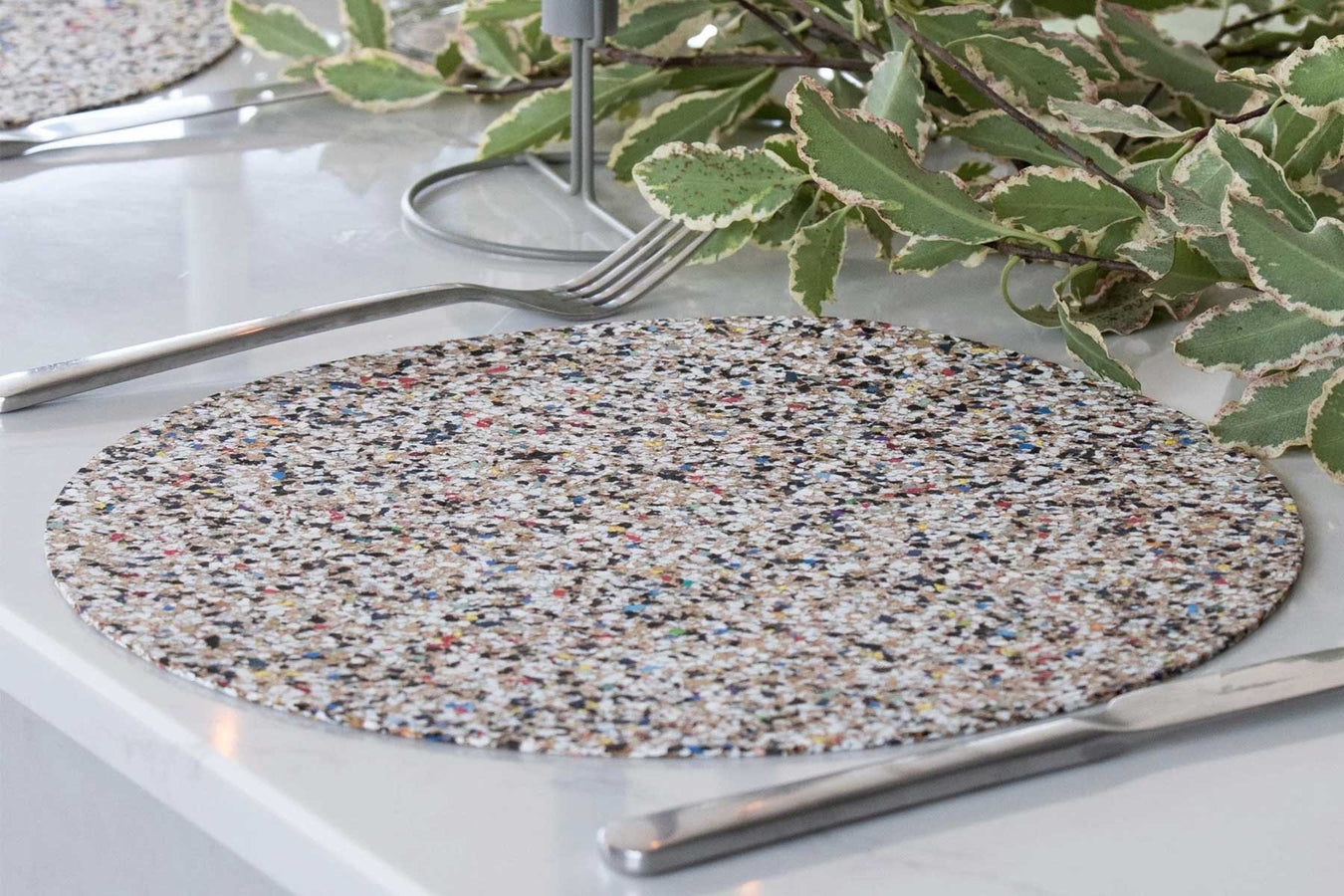 A speckled beachmat which has been recycled from beach plastics is laid on a table with cutlery and greenery.