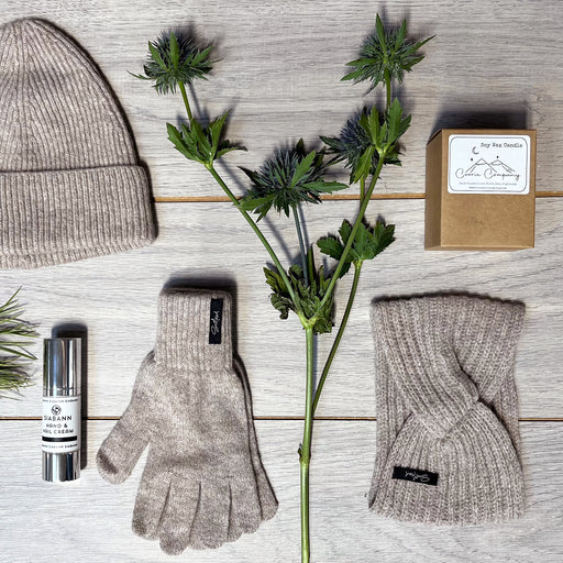 Knitted Hat, Gloves and Headband in stone laid on a wooden floor next to some thistles, a candle and Siabann hand cream 