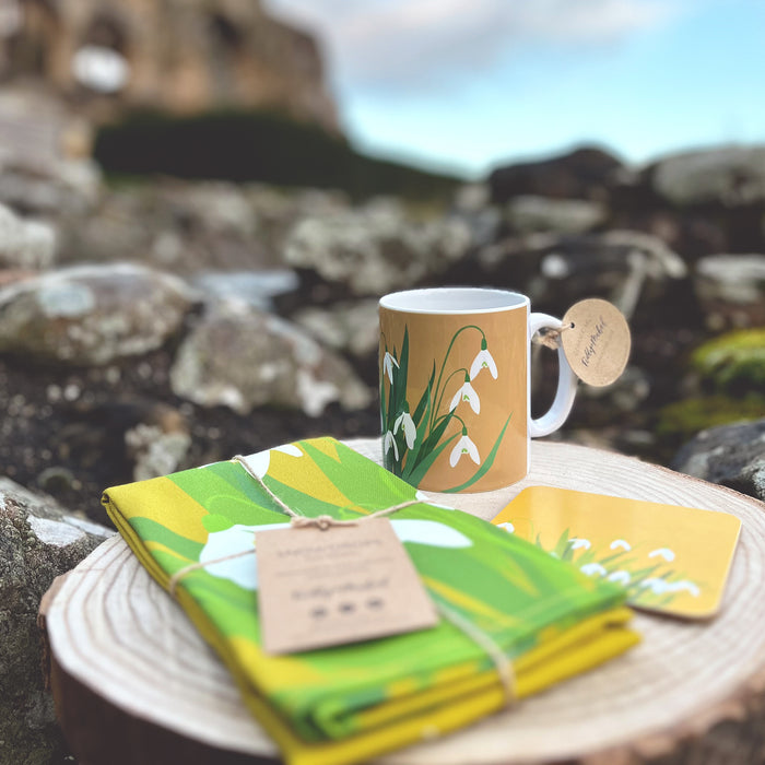 Snowdrop printed mug, teatowel and coaster placed on some rocks with blue skies and a old castle in the distance. 