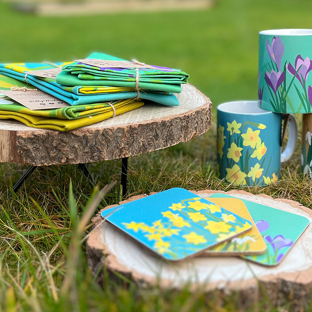 Selection of spring inspired kitchenware including tea towels, coaster and mugs. The items are arranged on a grassy scape and feature daffodil prints, crocus prints and snowdrop prints. 