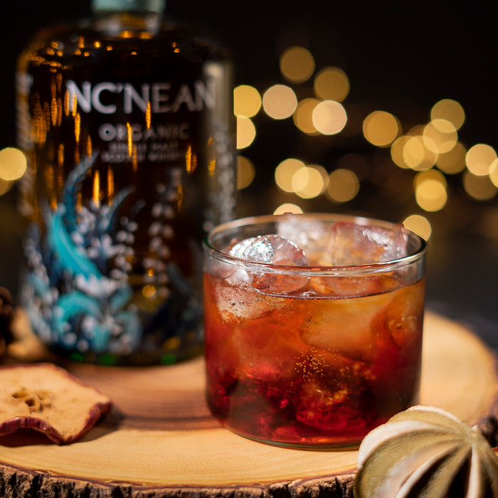 Bottle of NcNean Organic Single Malt Whisky next to a glass containing a whisky cocktail surrounded by festive dried fruit and fairy lights 