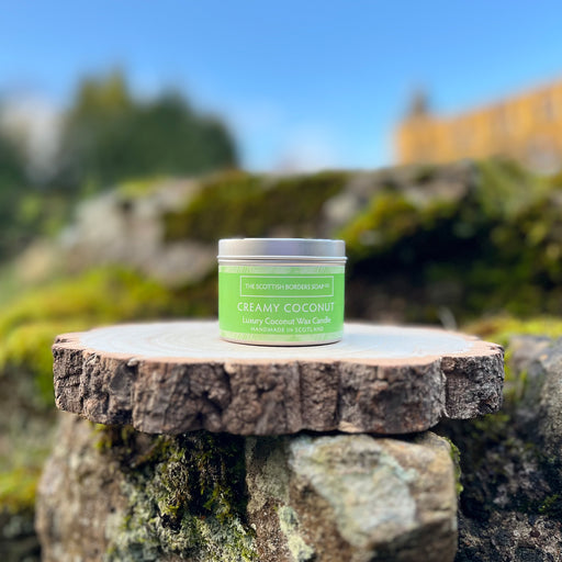 Silver tin with a green label containing the Creamy Coconut Candle sits on a piece of bark and rock with a sunny sky and castle in the background.