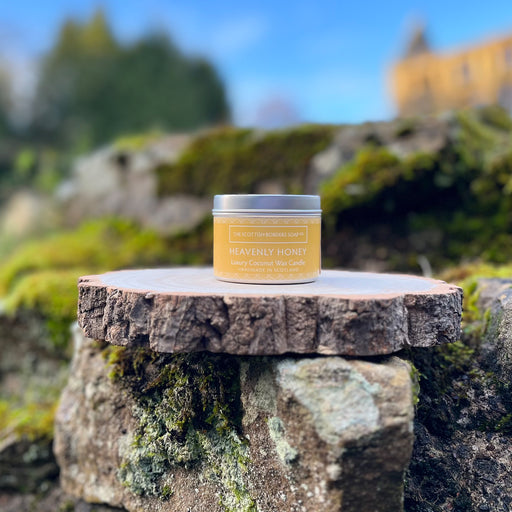 A silver Candle tin with a honey coloured label sits on a piece of bark and some rocks. In the background there is a blue sky and a blurred castle. 