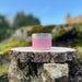 Silver Candle tin with a pink label sits on a piece of bark and rocks. The background shows blue skies and a castle in the distance. 