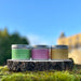 Trio of tins containing candles sit on a piece of bark and rock with a sunny sky and castle in the background.