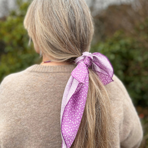 A purple twill tulip print scarf is tied around the long hair of a person who is standing in front of green trees.