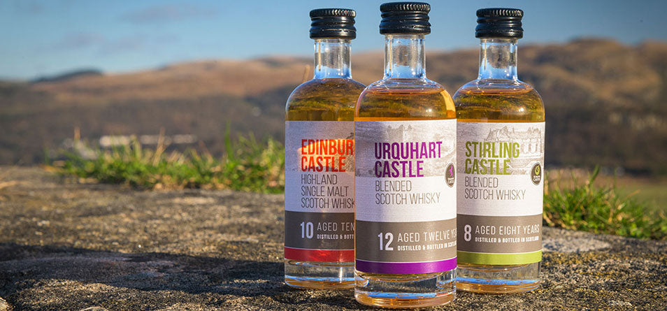 Trio of our exclusive Castle Whisky including Edinburgh, Urquhart and Stirling. The bottles are on a rugged path with hills and blue skies in the distance. 