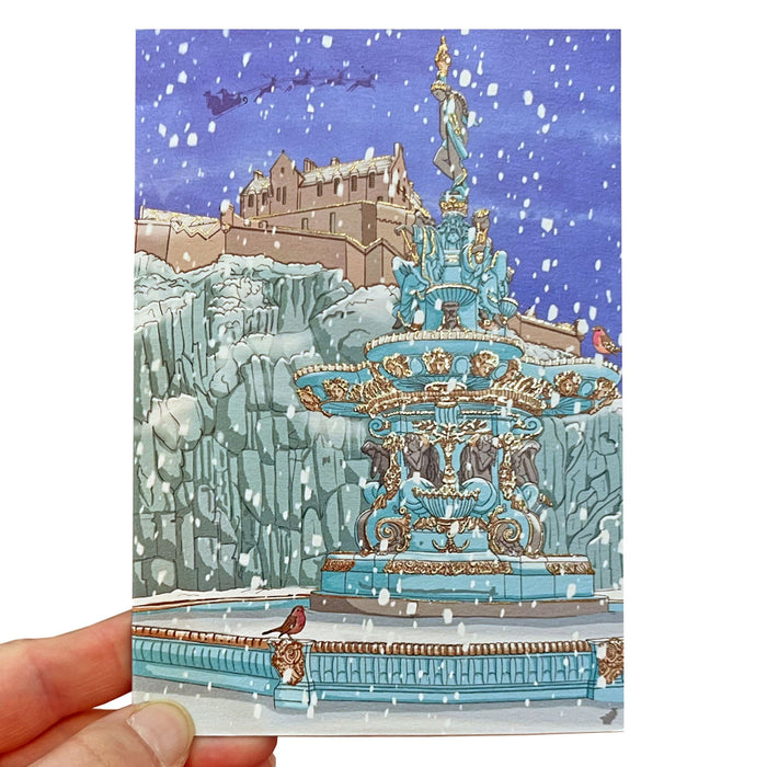 snowy illustration of edinburgh castle on christmas card front with robin perched on a large fountain in front of image. card is held by hand with fingers visible bottom left.  - Image