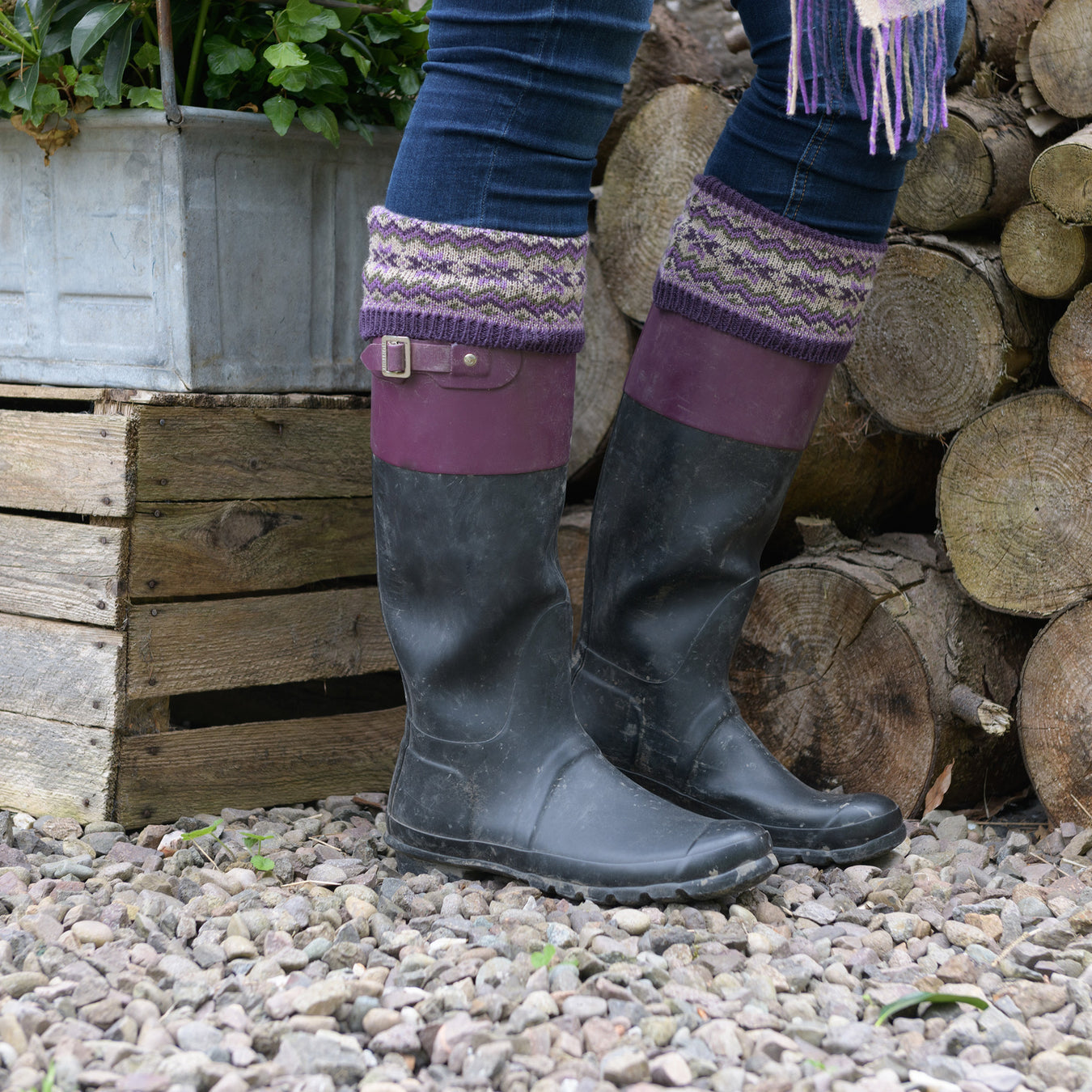 Person stands next to a pile of logs wearing a pair of black wellies and the Thistle coloured fairisle socks.