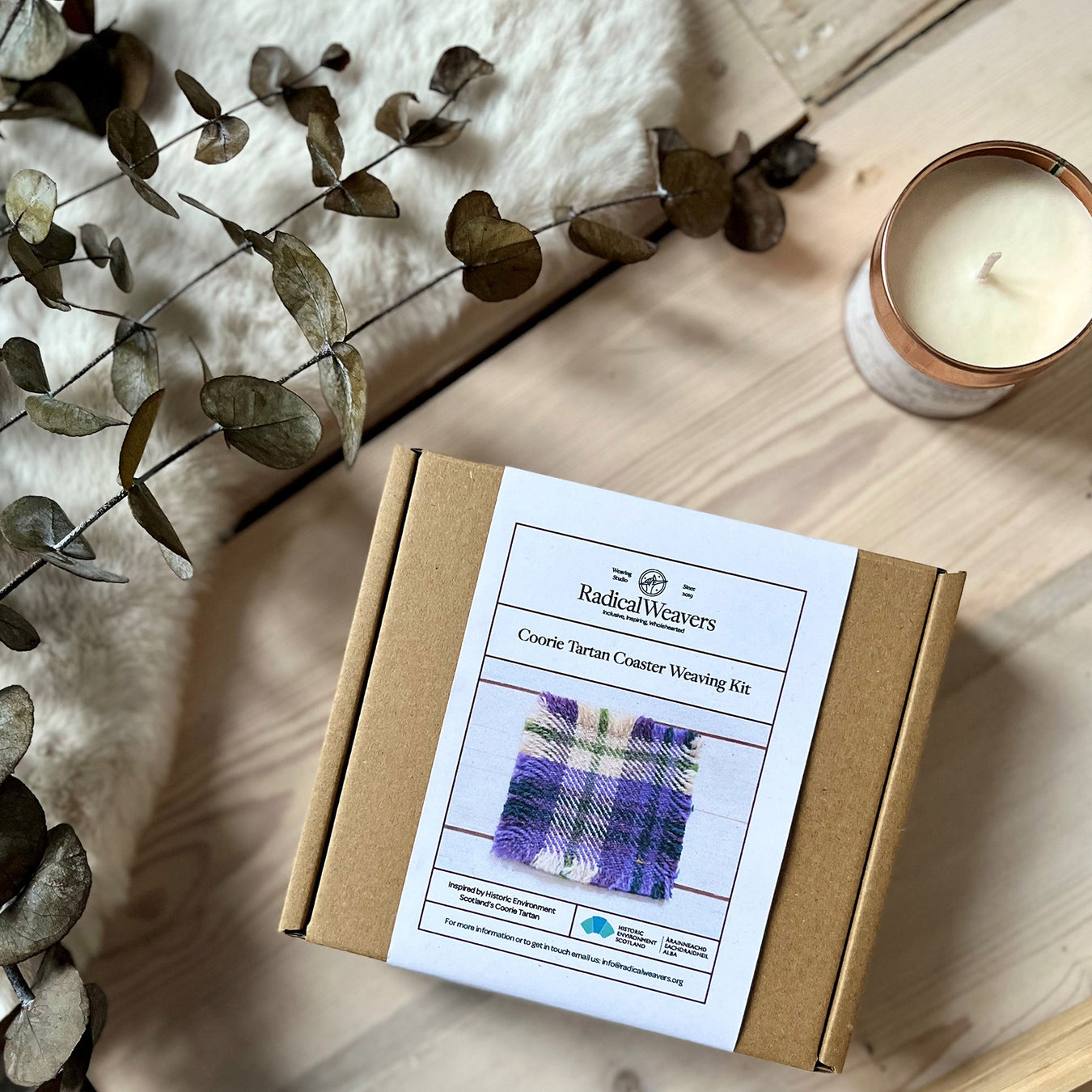 coorie tartan coaster weavign kit box on wooden floor next to a candle and rug with some foliage