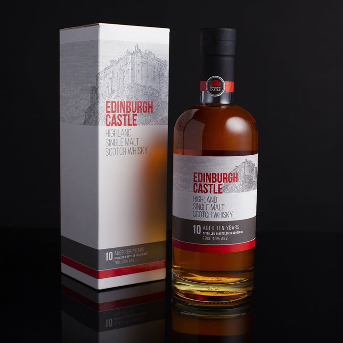 The official Edinburgh Whisky in a 70cl bottle is placed against a dark background next to its white presentation box. Both the box and bottle feature a sketch of Edinburgh castle in greyscale tone with the test 'Edinburgh Castle' in red. 