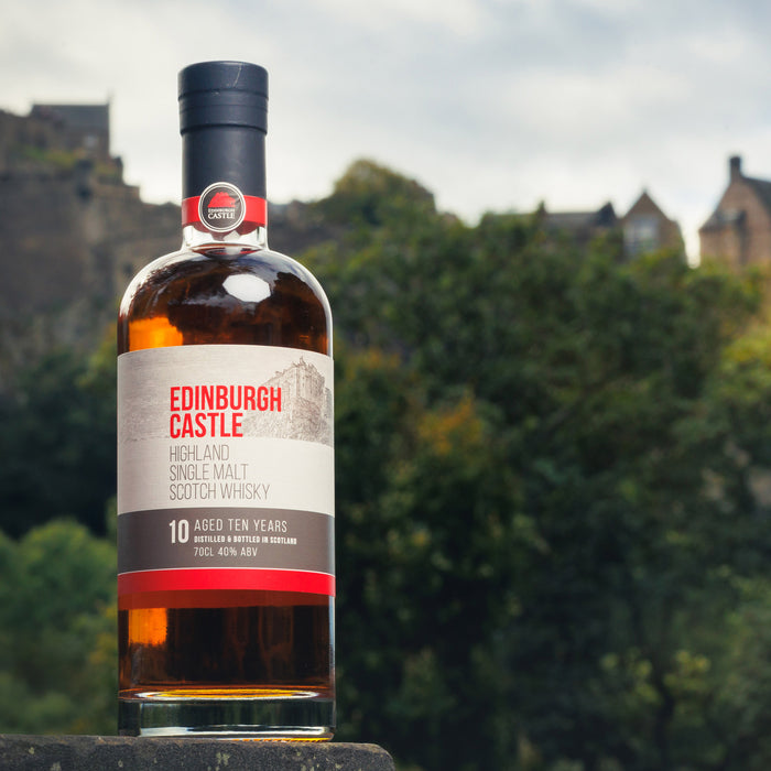 The official 70cl bottle of Edinburgh Castle Whisky. The label shows a sketch of the castle with the text in red reading 'Edinburgh Castle'. The bottle is sitting on a wall with the castle and trees in the background. 