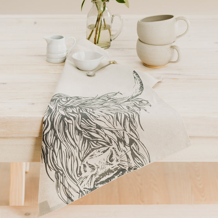 highland cow linen tea towel shown on table edge with cups and saucers