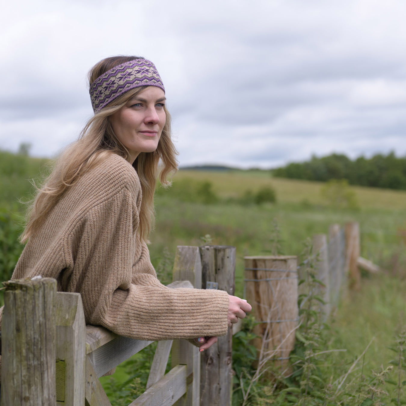 Person leaning on a fence overlooking the Scottish green countryside wears the Thistle coloured Fairisle headband. 