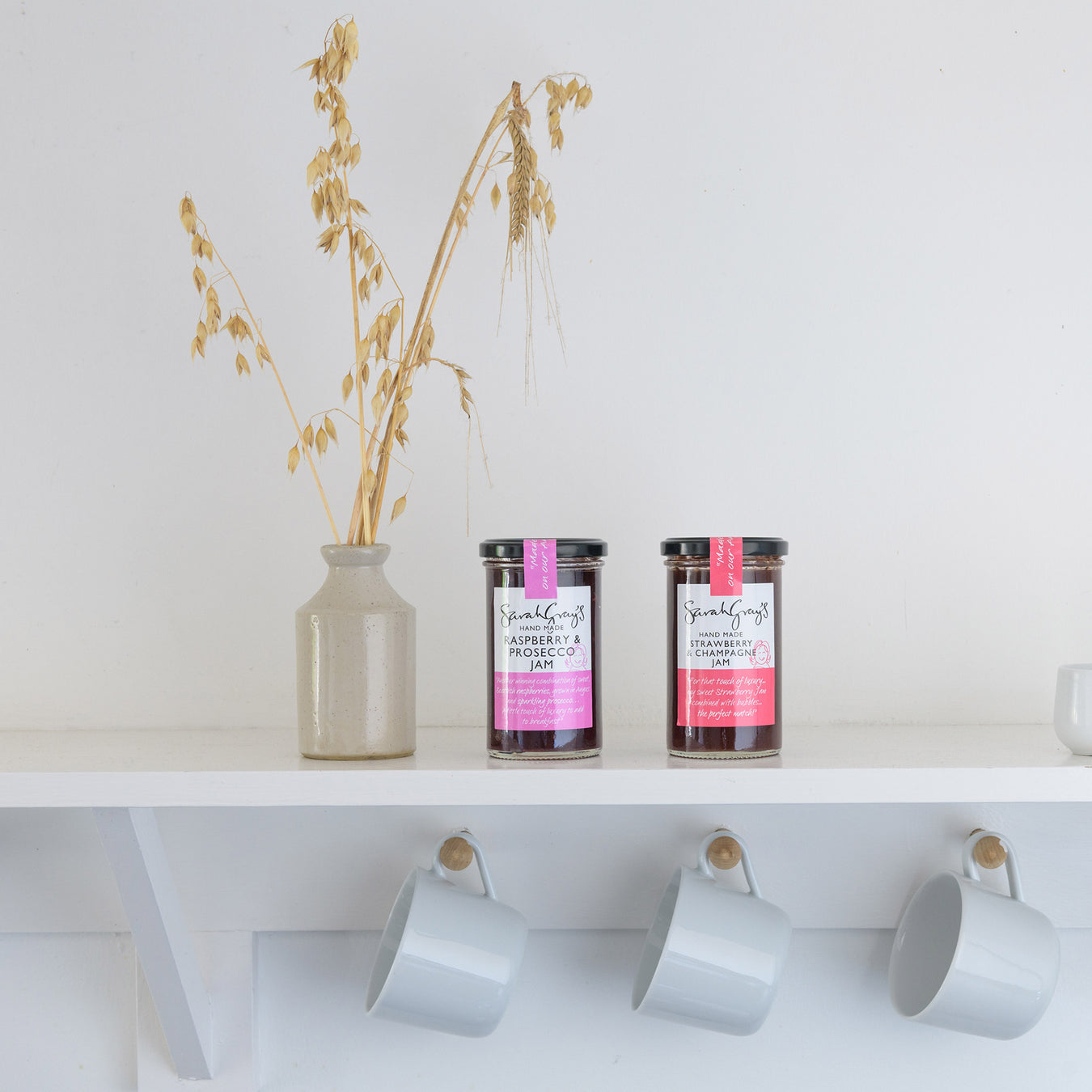 2 jam jars shown on a shelf next to a decorative jar with mugs hanging below shelf