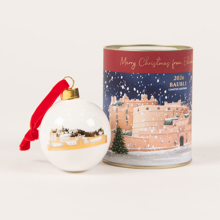A white Christmas ornament with a red ribbon and gold cap stands beside a cylindrical tin. The tin features a winter scene with a castle under falling snow and reads 'Merry Christmas from France' and '2026 BAUBLE LIMITED EDITION'. - Image