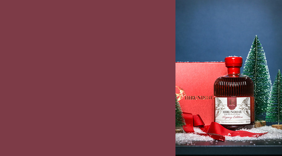 Hirundine Legacy Whisky bottle with a red cap on a snowy surface with Christmas trees in the background. - Image