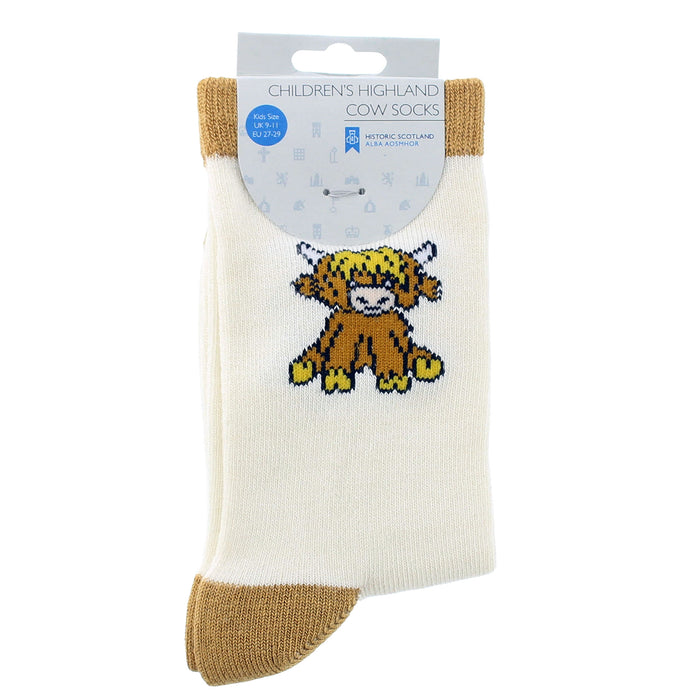 A pair of children's Highland cow socks in cream and tan, featuring a Highland cow graphic on the front, packaged on a white cardboard hanger. - Image
