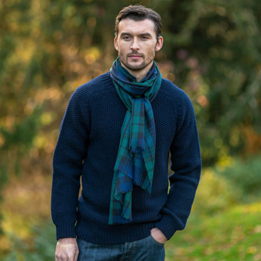 Man wearing a dark blue sweater and green and navy tartan scarf standing outdoors with blurred greenery in the background - Image