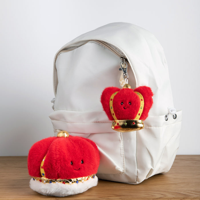 Two red plush toys with gold crowns attached to a white backpack on a wooden surface. - Image