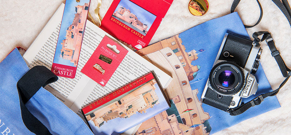 A styled flat-lay of Edinburgh Castle souvenir items on a light fabric background, including a blue tote bag, a small gold coin and magnet - Image
