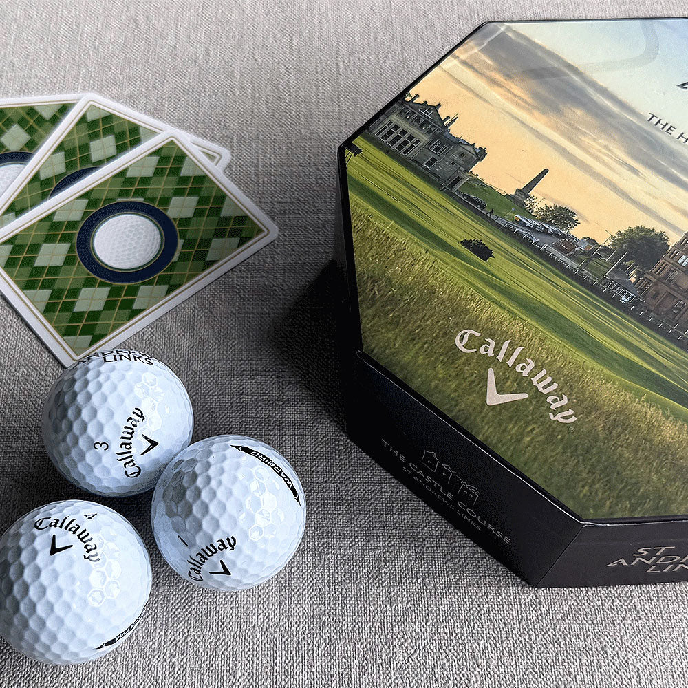 A Callaway St Andrews Links golf set displayed on a textured gray fabric surface, featuring three white golf balls, three green-and-white square cards, and a dark hexagonal tin box with a golf course illustration and Callaway branding. - Image