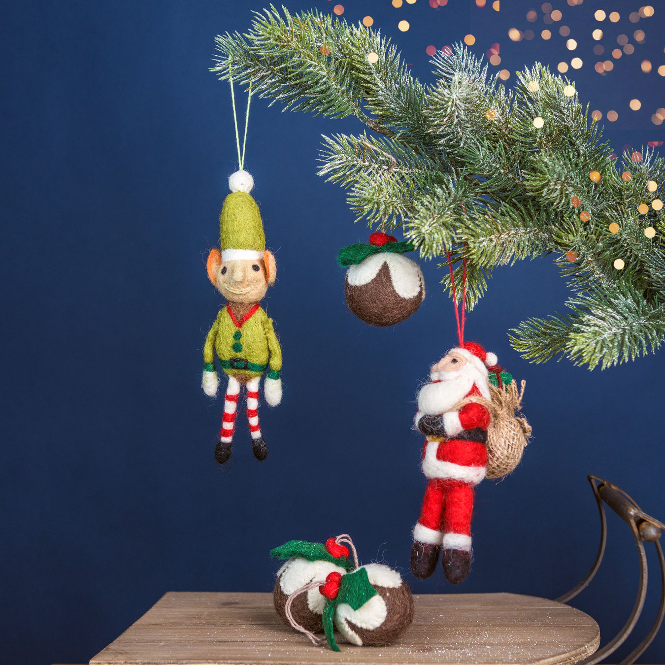 Christmas ornaments including a elf, pudding, and Santa Claus hanging from a tree branch against a dark blue background. - Image