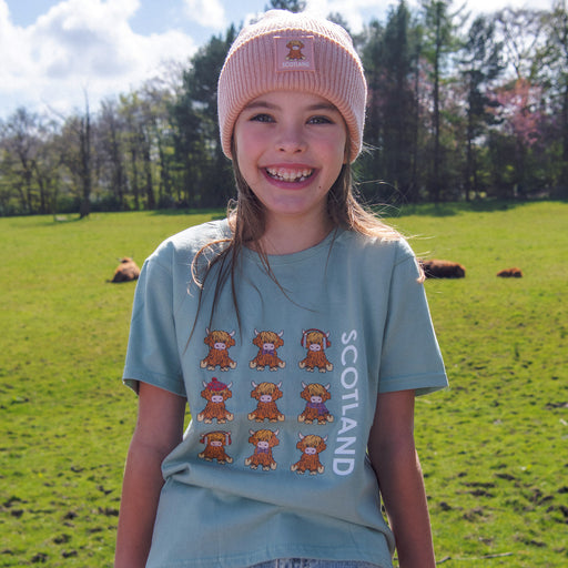 Young girl wearing a green t-shirt with cartoon highland cows and 'Scotland' text, standing in a grassy field. - Image