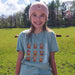 Young girl wearing a green t-shirt with cartoon highland cows and 'Scotland' text, standing in a grassy field. - Image