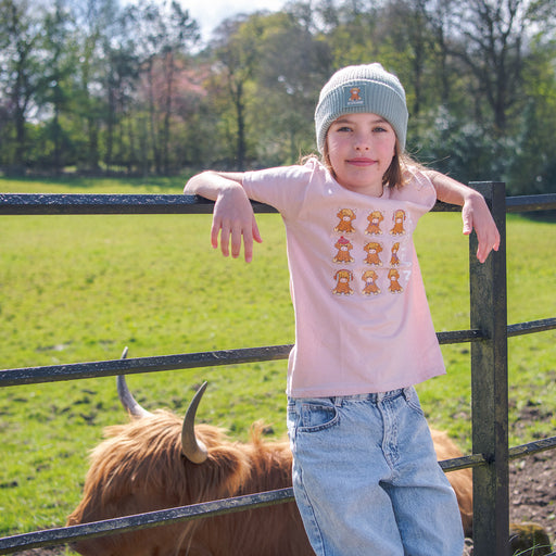 Child wearing a pink shirt with animal prints, standing next to a highland cow in a field. - Image