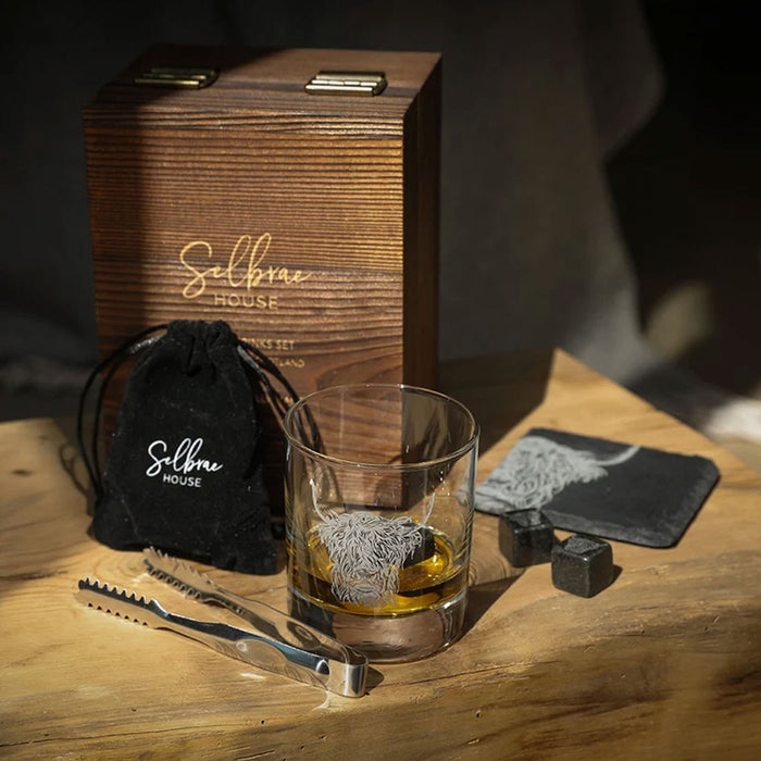 Whiskey glass, slate coaster both engraved with a Highland cow are placed with a wooden presentation box, velvet pouch, ice picker and whisky stones on a wooden surface. - Image
