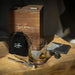 Whiskey glass, slate coaster both engraved with a Highland cow are placed with a wooden presentation box, velvet pouch, ice picker and whisky stones on a wooden surface. - Image