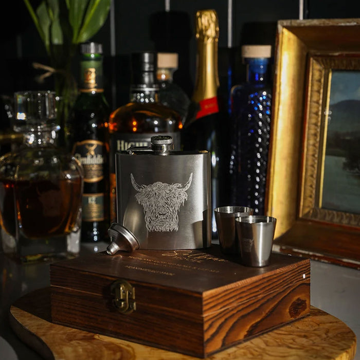 A silver stainless steel flask with an engraved Highland Cow design, presented on a wooden gift box with two small metal shot glasses. The set is styled on a wooden table with various liquor bottles and a gold picture frame in the background. - Image