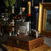 A silver stainless steel flask with an engraved Highland Cow design, presented on a wooden gift box with two small metal shot glasses. The set is styled on a wooden table with various liquor bottles and a gold picture frame in the background. - Image