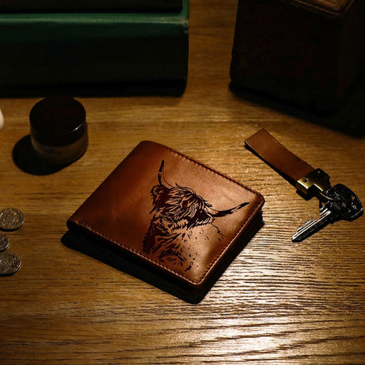 A brown leather bifold wallet with an engraved Highland cow design, resting on a wooden tabletop next to keys and coins. - Image