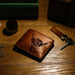 A brown leather bifold wallet with an engraved Highland cow design, resting on a wooden tabletop next to keys and coins. - Image