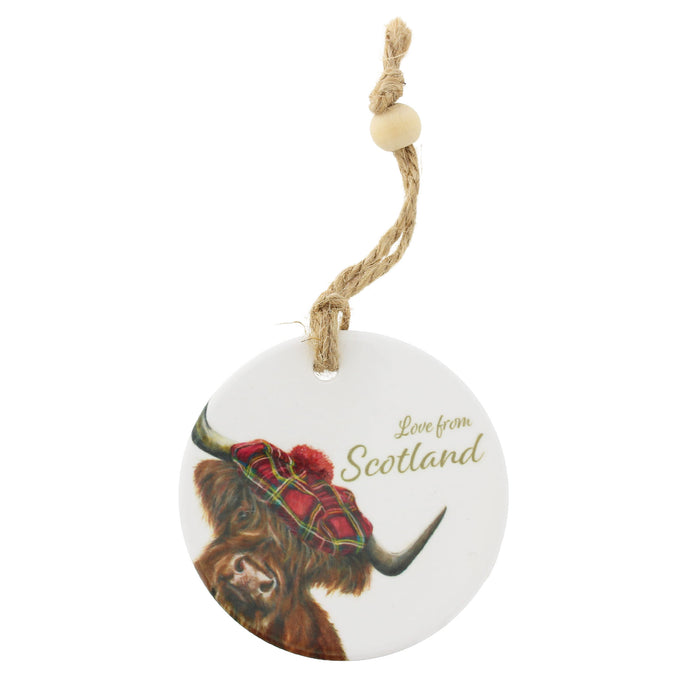 A round white ceramic ornament featuring a painted highland cow in a red tartan kilt with the text 'Love from Scotland' in gold, hanging from a natural jute rope with a wooden bead. - Image