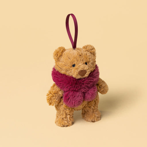 A small plush teddy bear decoration in warm tan with a fuzzy magenta scarf and a matching hanging ribbon, photographed on a plain warm beige background. - Image