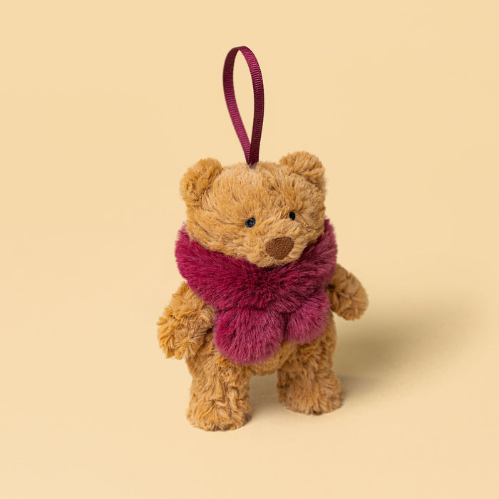 A small plush teddy bear decoration in warm tan with a fuzzy magenta scarf and a matching hanging ribbon, photographed on a plain warm beige background. - Image