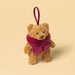 A small plush teddy bear decoration in warm tan with a fuzzy magenta scarf and a matching hanging ribbon, photographed on a plain warm beige background. - Image