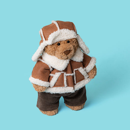 A plush teddy bear dressed in a brown aviator-style jacket and matching hat with white faux-fur trim, standing against a solid light blue background. - Image