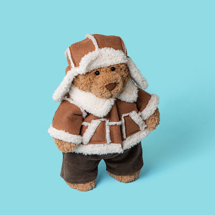 A plush teddy bear dressed in a brown aviator-style jacket and matching hat with white faux-fur trim, standing against a solid light blue background. - Image