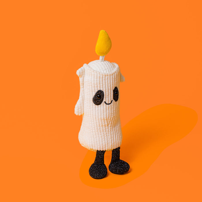 A cute, novelty plush toy shaped like a candle with a ribbed white body, black embroidered face, and black glittery feet, standing against a solid orange background. - Image